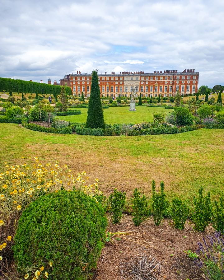 Hampton Court Palace London Less Touristy Places in London for Return Visitors A manicured garden in London with green trees and bushes in circular patterns. Behind is Hampton Court Palace which is a large red and white rectangular building with lots of little windows in it and above is a cloudy sky. Less Touristy Places in London for Return Visitors