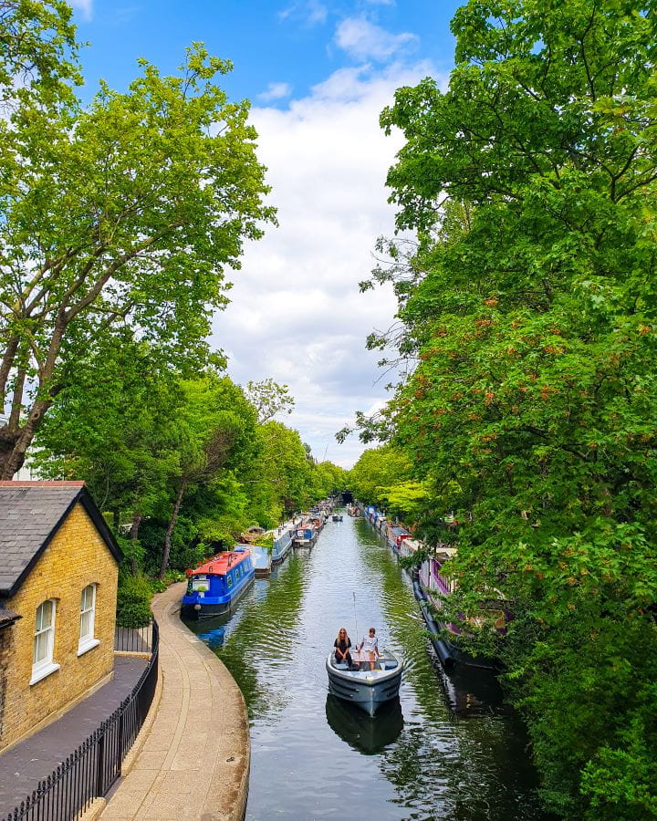 Little Venice London Less Touristy Places in London for Return Visitors A canal running down the middle of the photo with canal boats moored on either side and a boat sailing down the middle. There's a little building on one side and lots of trees in Little Venice in London. Less Touristy Places in London for Return Visitors