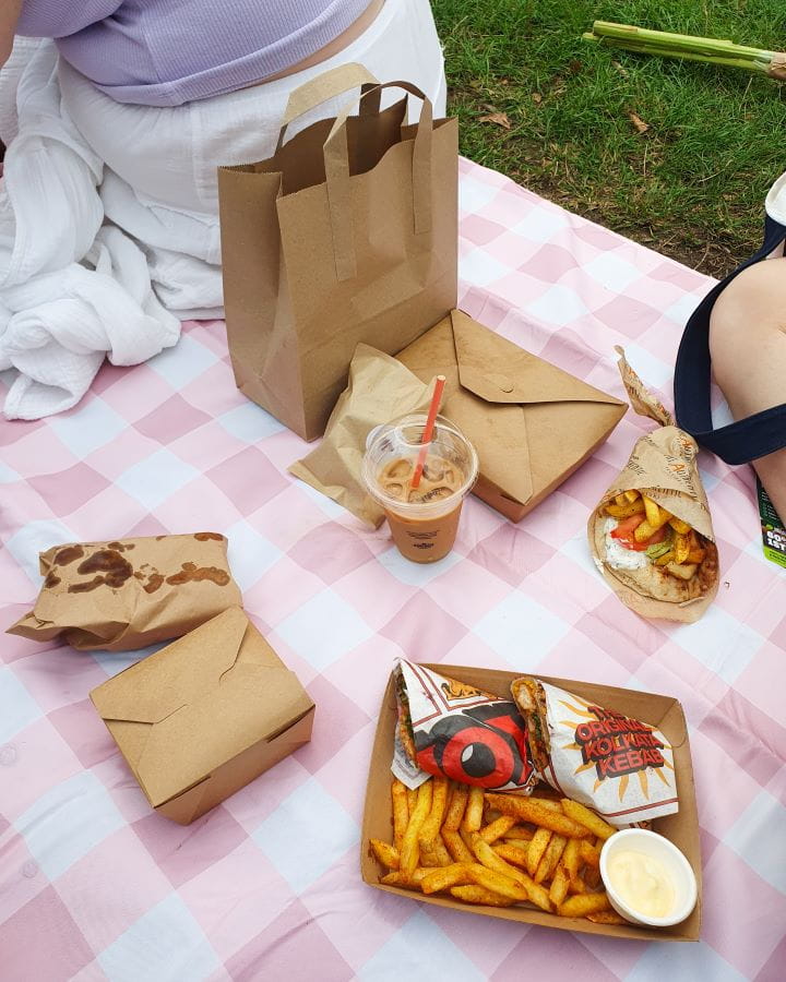 Broadway Market London Fields London Less Touristy Places in London for Return Visitors Some food on a pink and white picnic blanket in London Fields in London. There's some paper containers and bags with food and a tray with a wrap and some chips in it. Less Touristy Places in London for Return Visitors