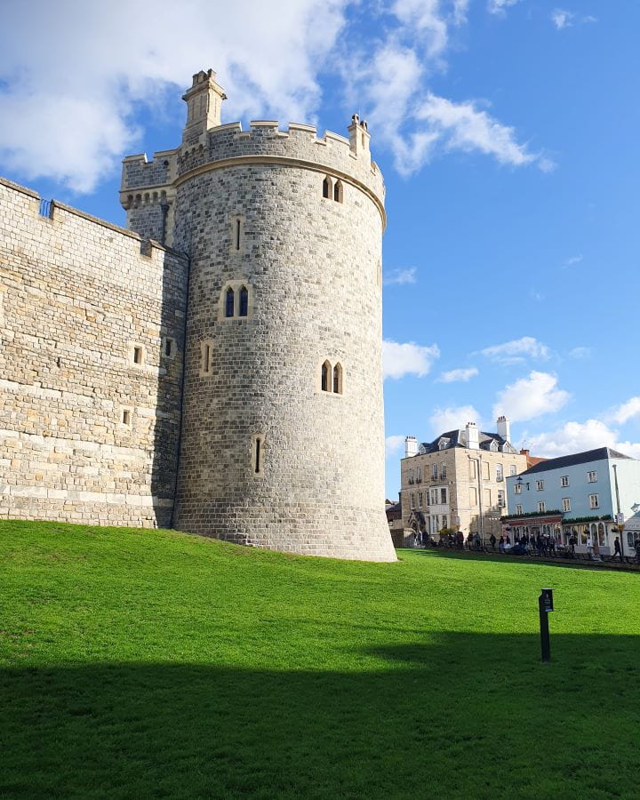 Windsor Castle Windsor London Less Touristy Places in London for Return Visitors Windsor Castle in London. There's a big green patch of grass with part of the stone castle wall and turret behind. There's little windows in the castle turret and on the right of it is some little blue and beige coloured shops. Less Touristy Places in London for Return Visitors