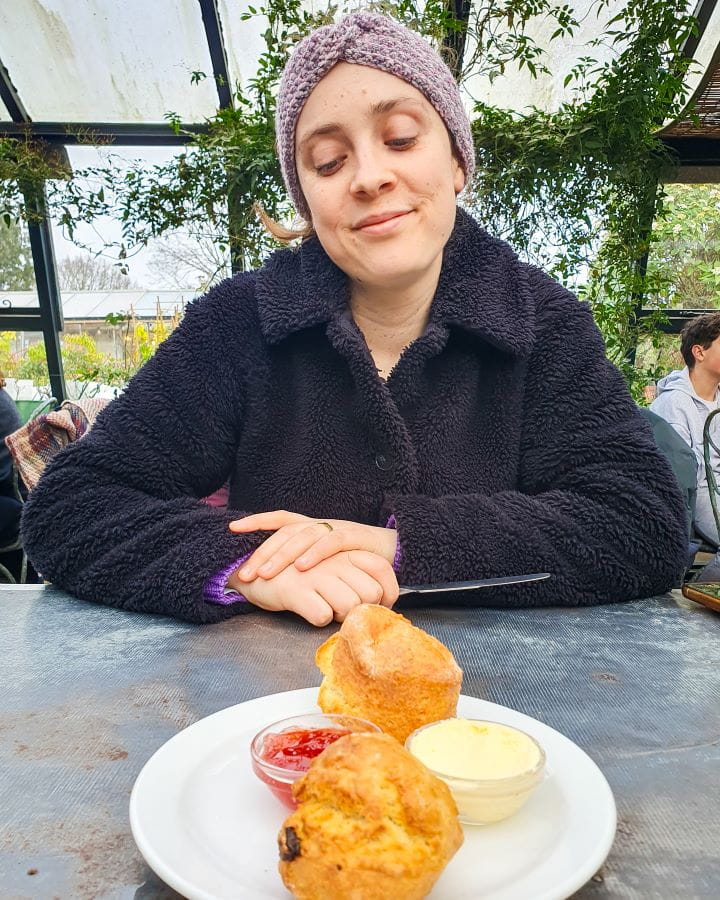 Richmond Petersham Nurseries London Less Touristy Places in London for Return Visitors Krissie wearing a black fluffy coat and looking down at a plate of scones on the table at Petersham Nurseries in London. There's two scones on the plate with a little bowl of jam and another one of cream. Behind her is glass panels with green vines all over them. Less Touristy Places in London for Return Visitors