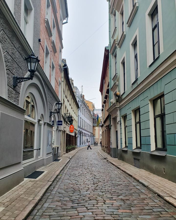 A cobbled street in Riga, Latvia. The street is narrow and there's coloured buildings on either side. There's barely anyone on the street and the sky is overcast. How to Make the Most of Riga In One Day