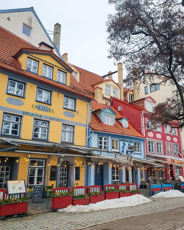 How to Make the Most of Riga In One Day. Three coloured buildings in Līvu Square, Riga, Latvia. The building on the left is yellow and blue, the middle one is blue with an orange roof, and the right one is red and pink. There's outdoor seating outside with fairy lights, and a tree on the right.