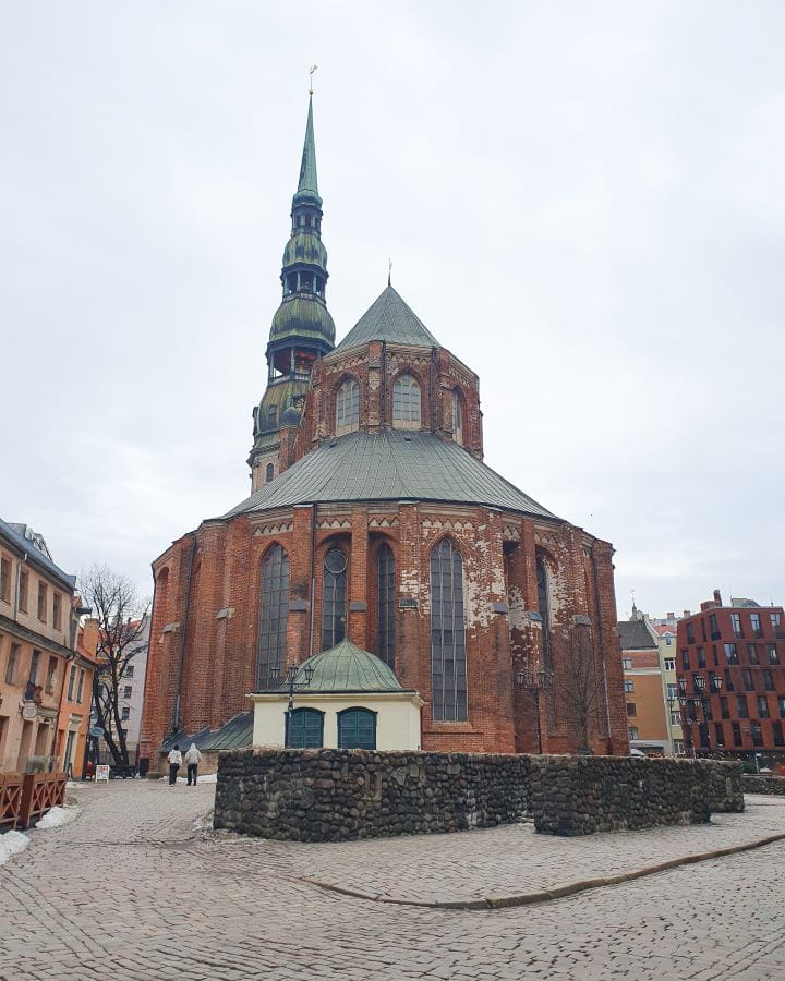 A big and round orange church with a green roof in Riga, Latvia. There's a tall spire behind it and a brick path around it with other buildings too. How to Make the Most of Riga In One Day