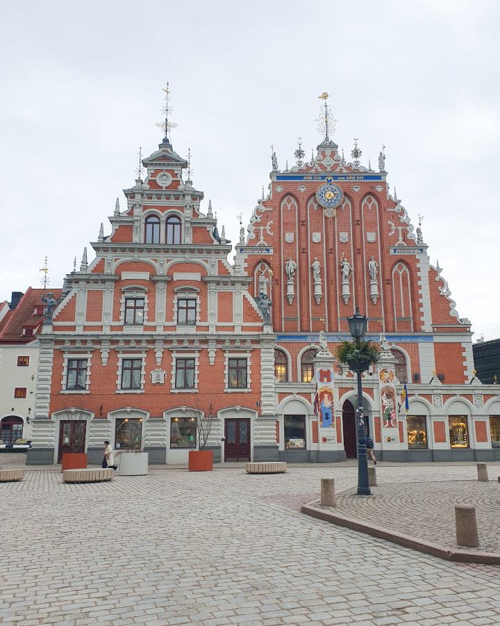 Two fancy buildings in Riga, Latvia. They're orange and white, with points coming off them and windows on the front. In front of them is a paved path with some seats and lamp posts and there's no one around. How to Make the Most of Riga In One Day