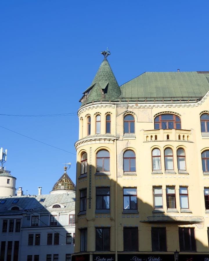 A beige house with a green roof that has a turret with a small cat statue on top of it in Riga, Latvia. There's lots of windows on the building and there's a white building behind it too. How to Make the Most of Riga In One Day