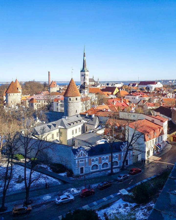 A view over the city of Tallinn, Estonia. There's the old town, with orange rooves, turrets and church spires. The sky above is clear and blue and in the foreground is a road with cars on it. How to Make the Most of Riga In One Day