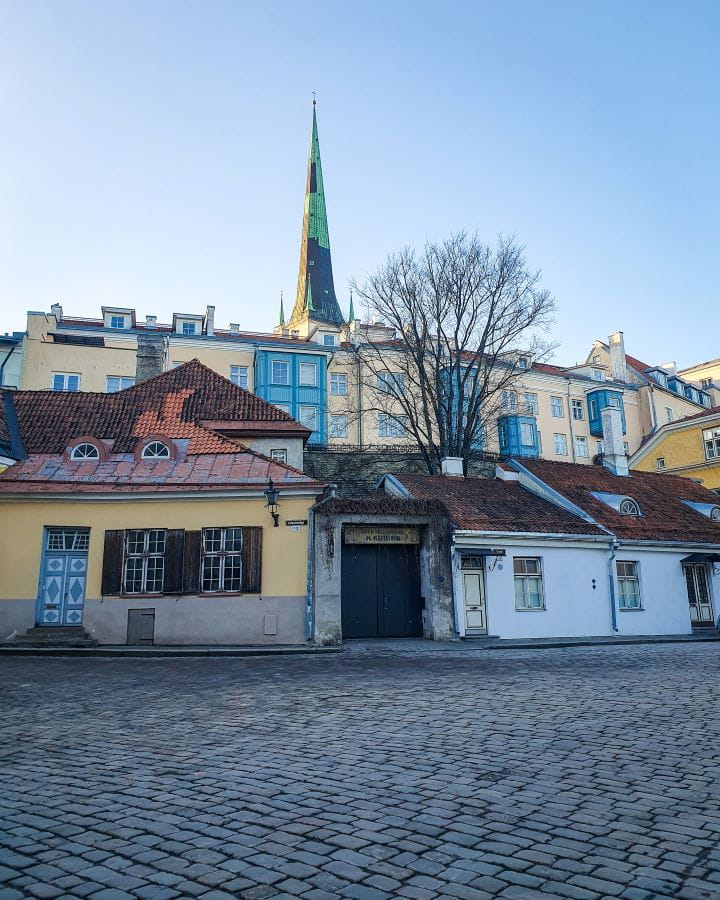 Tallinn Estonia How To Do Helsinki and Tallinn In a Weekend How To Do Helsinki and Tallinn In a Weekend. An empty street in Tallinn, Estonia. There's white, yellow and beige houses with red roofs. There's a bare tree in the middle and a green church spire behind it all.