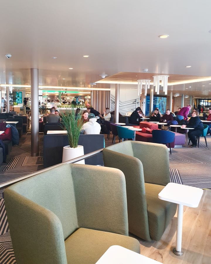 Tallink How To Do Helsinki and Tallinn In a Weekend A seating area on a Tallink ship. In the foreground are two big soft green chairs with white tables. Behind is another big seating area with tables in front of all the chairs, and a café behind them all. How To Do Helsinki and Tallinn In a Weekend