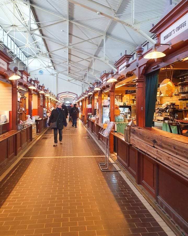 Old Market Hall Helsinki Finland How To Do Helsinki and Tallinn In a Weekend Inside the old market hall in Helsinki, Finland. There's brown shop windows on either side of a path, with each one selling delicious food and drink. There's people walking down the path and above is a white roof. How To Do Helsinki and Tallinn In a Weekend