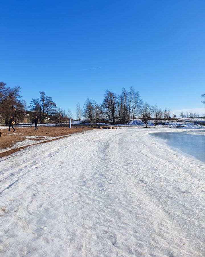 Helsinki Finland How To Do Helsinki and Tallinn In a Weekend On a beach on an island in Helsinki, Finland. You can see sand on the left and the rest of the beach has snow on it. On the right is some water and behind are trees and paths with people walking around. How To Do Helsinki and Tallinn In a Weekend