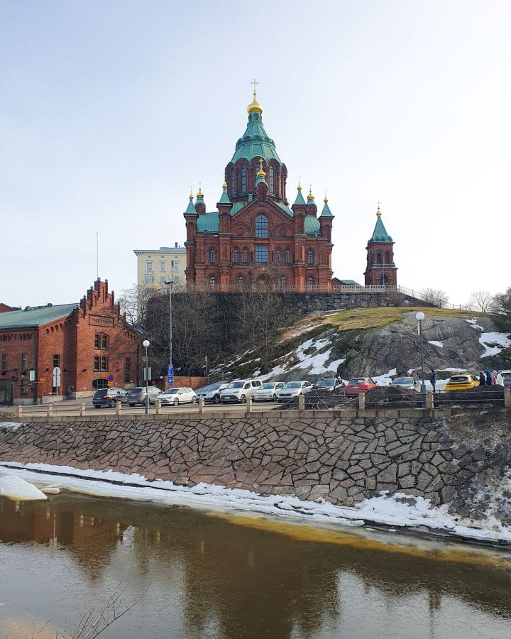 Uspenski Cathedral Helsinki Finland How To Do Helsinki and Tallinn In a Weekend A brown Cathedral with a pointy green roof on a hill in Helsinki, Finland. Under the hill is a carpark with cars parked in it and a brown building on the left. The Cathedral has lots of pointy green turrets and a big dome in the middle with a gold cross on top. How To Do Helsinki and Tallinn In a Weekend