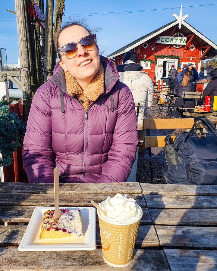 Café Regatta Helsinki Finland How To Do Helsinki and Tallinn In a Weekend Krissie in a purple puffer jacket sitting at a wooden table in Helsinki, Finland. On the table is a blueberry cake and a hot chocolate with whipped cream. Behind her are more people and a little red wooden hut that says Regatta on it. How To Do Helsinki and Tallinn In a Weekend