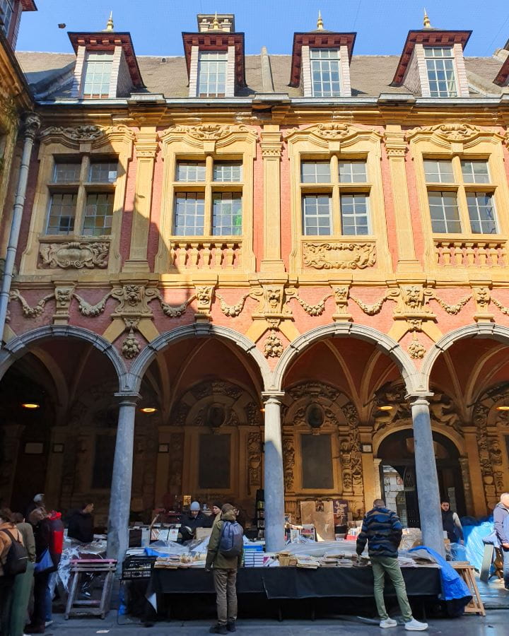 Inside a courtyard in Lille, France. It's a beautiful yellow and orange building with grey archways. Underneath are tables with old books and paintings and lots of people around looking at it all. Above are eight windows and the sky above is blue. How To Enjoy Lille in a Day