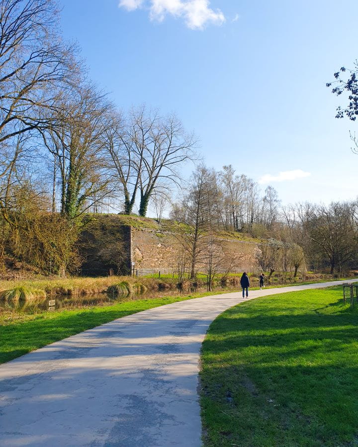 A path with green grass on either sides with a tall brick wall on the far left next to a small river. There's trees around and other people on the path ahead beside the Citadelle in Lille, France. How To Enjoy Lille in a Day