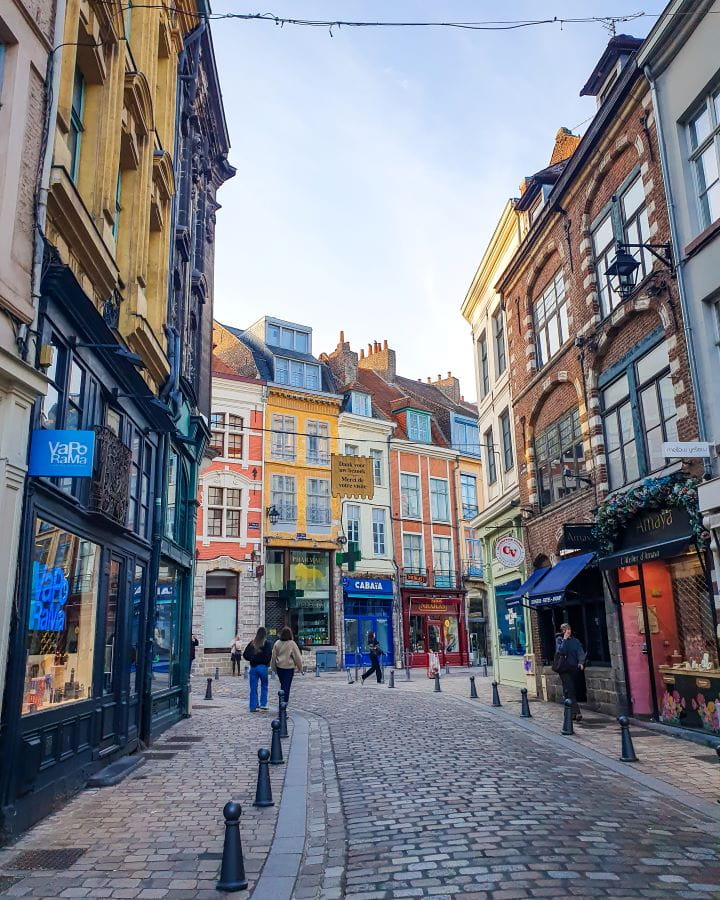 A street in Lille, France with cobbled paths and a road running between them. There's 3 or 4 storey buildings on either side of the road and coloured buildings at the end in orange, yellow and white. There's some people walking around and the sky is going into sunset. How To Enjoy Lille in a Day