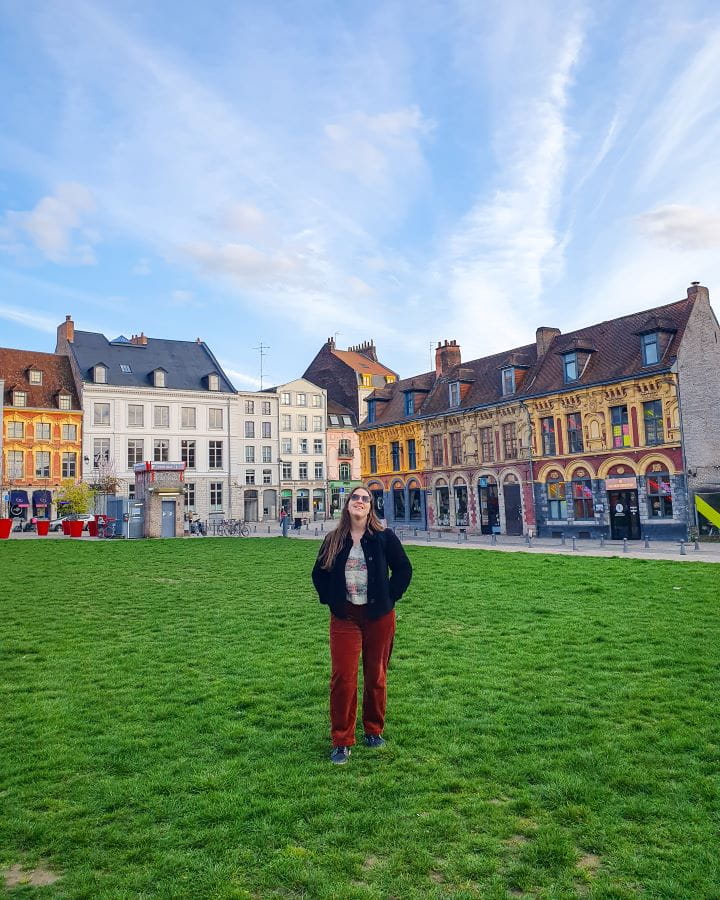 How To Enjoy Lille in a Day. Katherine wearing orange pants, a black coat and standing in the middle of a big patch of grass in Lille, France. Behind her are lots of coloured European buildings with triangular roofs. The sky is blue with wispy clouds above it all.
