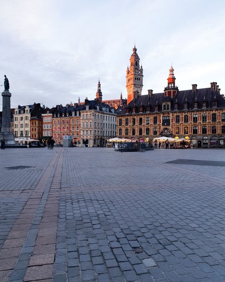 A town square in Lille, France. There's beautiful European buildings coloured in gold, white, orange and red. Behind is a big church tower and on the left is a tall monument. The square is pretty empty, there's some outdoor seating and the photo is taken at sunset so it's getting dark. How To Enjoy Lille in a Day
