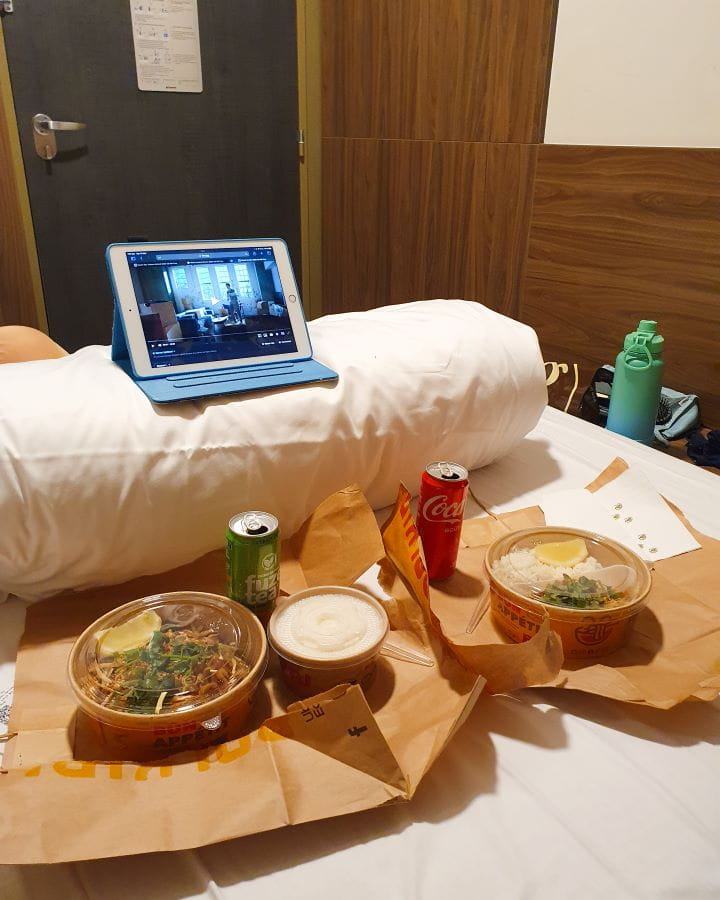 Inside our hotel room in Lille, France. The photo is taken on a white bed sheet with two paper bags and two takeaway containers on top with thai food in them. There's also two cans of soft drink and a coloured water bottle on the side. There's a white rolled up doona with a blue ipad on top with a show playing on it. Behind the bed is a wooden looking wall and a blue door. How To Enjoy Lille in a Day