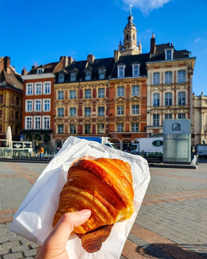 In the main square in Lille, France. The main part of the photo is a hand holding a white paper bag that says "The French Bastards" on it with a big beautiful golden croissant on top. Behind the hand are the coloured buildings of the main square and there's a bright blue sky above. How To Enjoy Lille in a Day