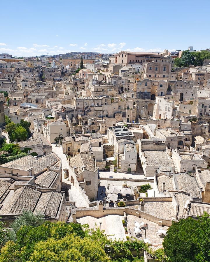 Matera Italy Three Unique Day Trips From Bari Without a Car A view over Matera in Italy. In the foreground are green trees and behind it are the cream coloured buildings and white small black windows of the city. Three Unique Day Trips From Bari Without a Car
