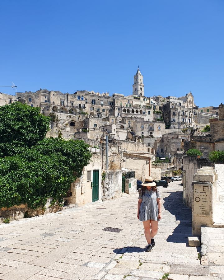 Matera Italy Three Unique Day Trips From Bari Without a Car Katherine walking up a white tiled street in Matera, Italy. On the left is a big green bush and behind are lots of cream buildings with a church spire coming up from the middle. Three Unique Day Trips From Bari Without a Car
