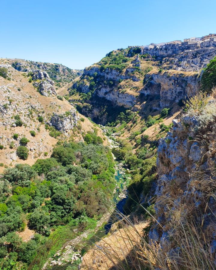 Matera Italy Three Unique Day Trips From Bari Without a Car A valley with a thin blue river going through the middle. There's lots of green bushes and trees as well as brown grass and rocks in the valley and on the top right you can see some of the cream buildings of Matera, Italy. Three Unique Day Trips From Bari Without a Car