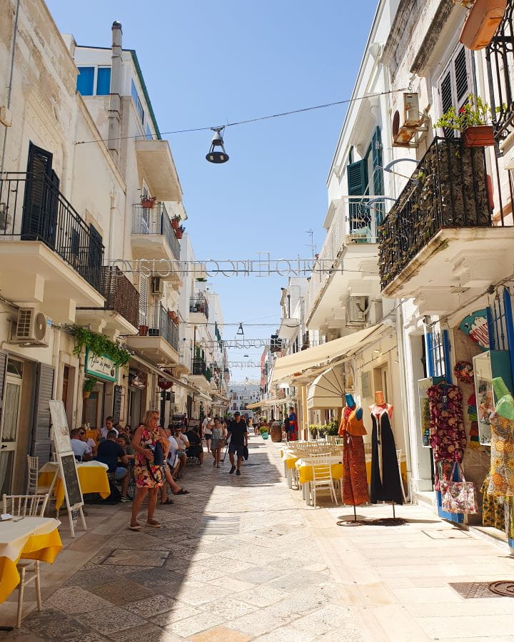 Polignano a Mare Italy Three Unique Day Trips From Bari Without a Car A street in Polignano A Mare in Italy. The street has white tiles on the ground and white buildings on either side. Between the buildings are light up lyrics and lights above and on the streets are tables and displays from the shops and restaurants. Three Unique Day Trips From Bari Without a Car