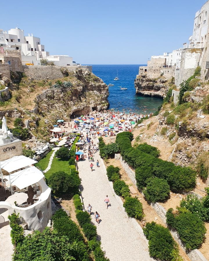 Lama Monachile beach Polignano a Mare Italy Three Unique Day Trips From Bari Without a Car A white path with green trees on either side leading up to Lama Monachile beach in Polignano A Mare, Italy. There's lots of people and beach umbrellas on the beach with blue water behind it with boats on it.. There's rock cliffs on either side of the water with white buildings on them. Three Unique Day Trips From Bari Without a Car