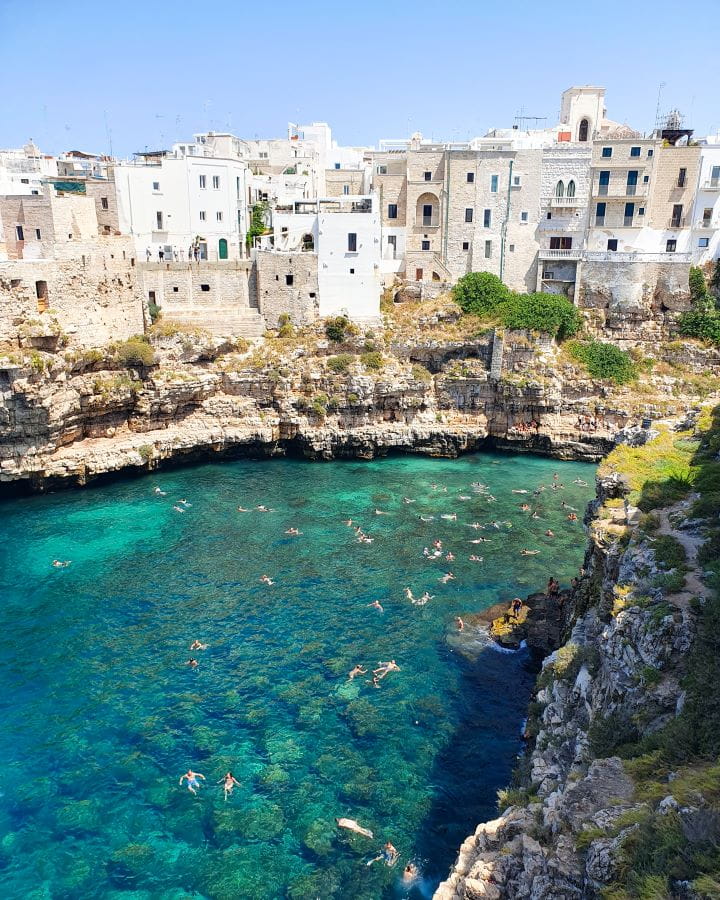 Lama Monachile beach Polignano a Mare Italy Three Unique Day Trips From Bari Without a Car A view over the water and cliffs in Polignano A Mare in Italy. In the foreground is clear blue water with people floating in it. Behind them is a rocky cliff and on the edge of the rocks are lots of white buildings with little black windows in them. Three Unique Day Trips From Bari Without a Car
