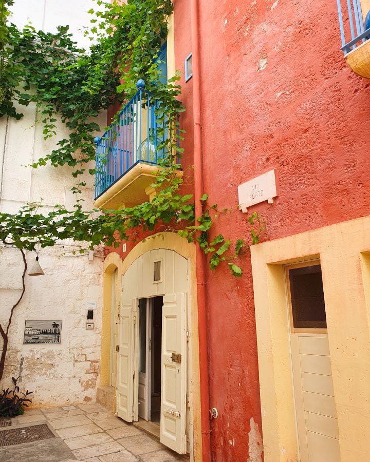 Polignano a Mare Italy Three Unique Day Trips From Bari Without a Car In the old town in Polignano A Mare in Italy. There's a tall red wall with yellow doors and windows and blue balconies. There's also green vines winding around the walls. Three Unique Day Trips From Bari Without a Car