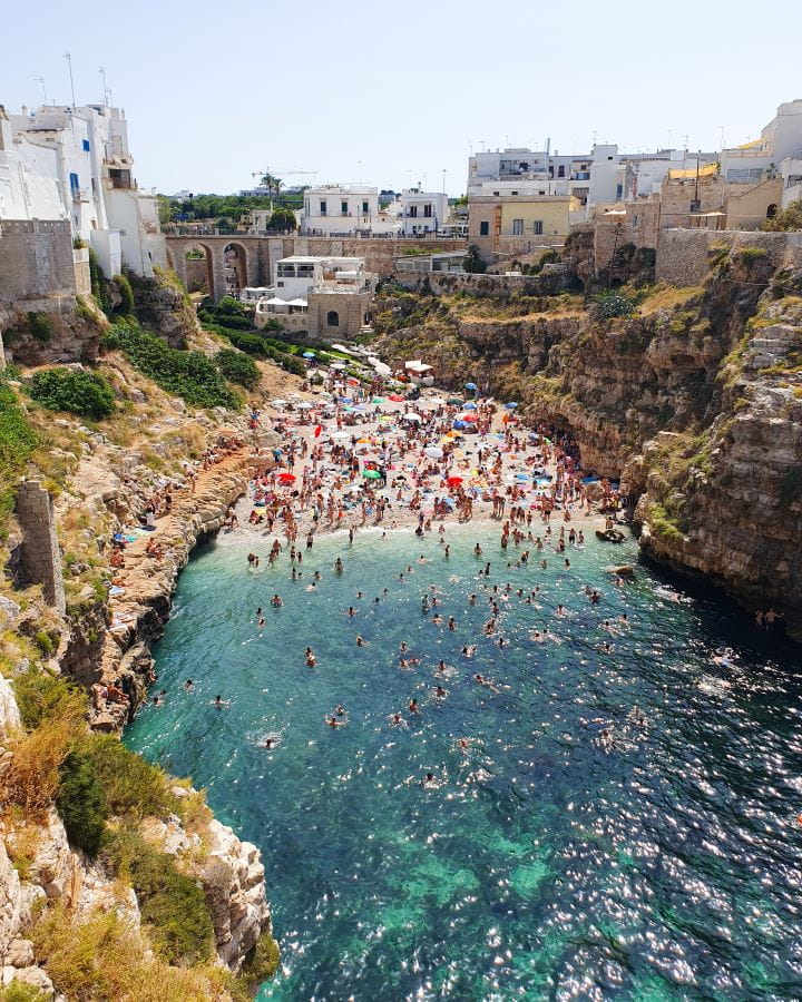 Lama Monachile beach Polignano a Mare Italy Three Unique Day Trips From Bari Without a Car Three Unique Day Trips From Bari Without a Car. Lama Monachile beach in Polignano A Mare nestled within a cove with rocks on either side. The beach and water are packed with people and there's white buildings on the cliffs above along with green grass and trees.