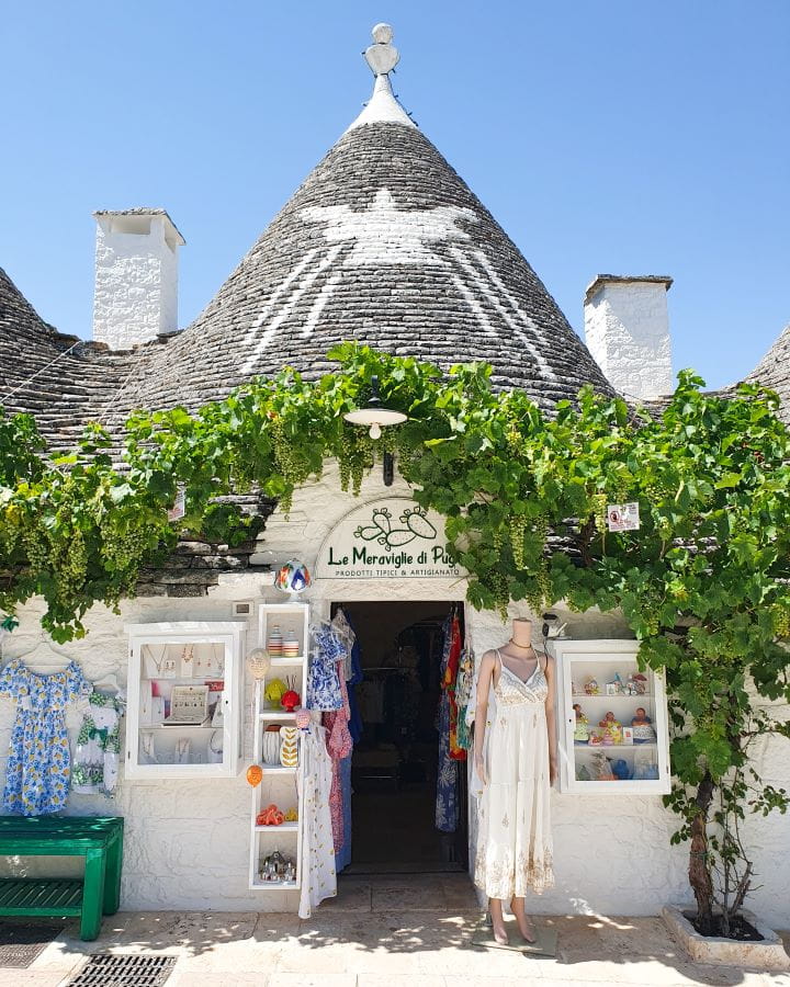 Alberobello Italy Three Unique Day Trips From Bari Without a Car A white trullo with a grey roof that also has a white shape painted on it in Alberobello, Italy. There's a big green vine running along the top of the doorway of the trullo and out the front is clothes and a mannequin displaying stuff from the shop inside. Three Unique Day Trips From Bari Without a Car