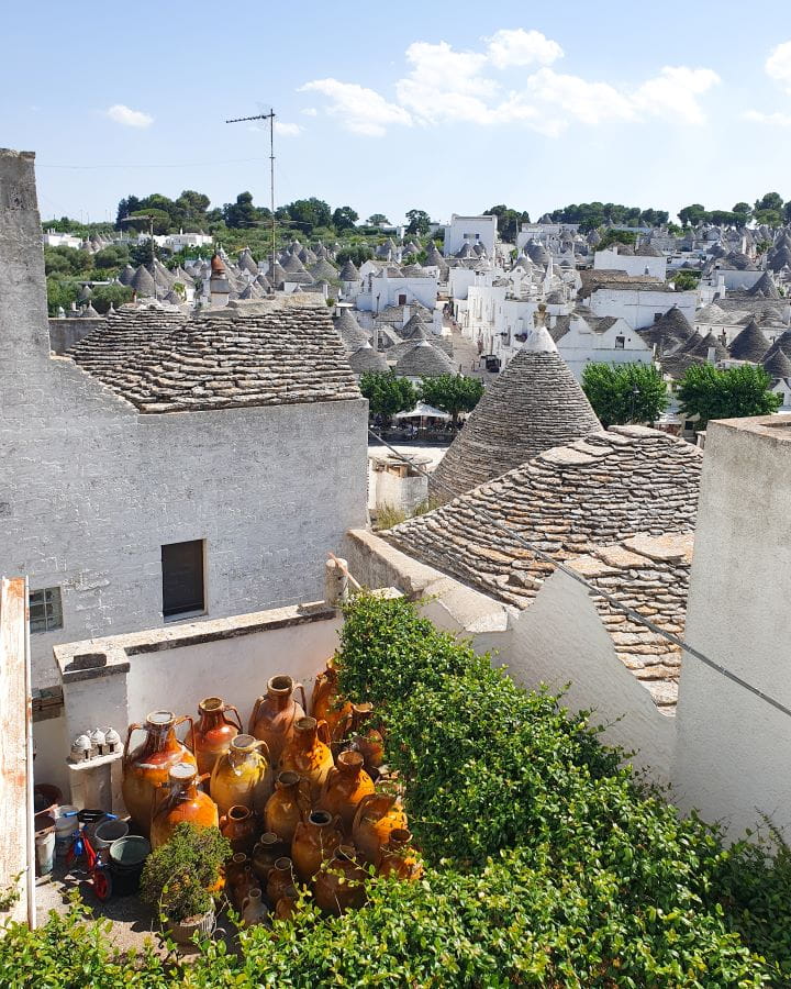 Alberobello Italy Three Unique Day Trips From Bari Without a Car A view over the trulli of Alberobello, Italy. In the foreground is a big green plant and some brown jugs. Behind that are lots of cone shaped trulli with white buildings and grey rooves. Three Unique Day Trips From Bari Without a Car