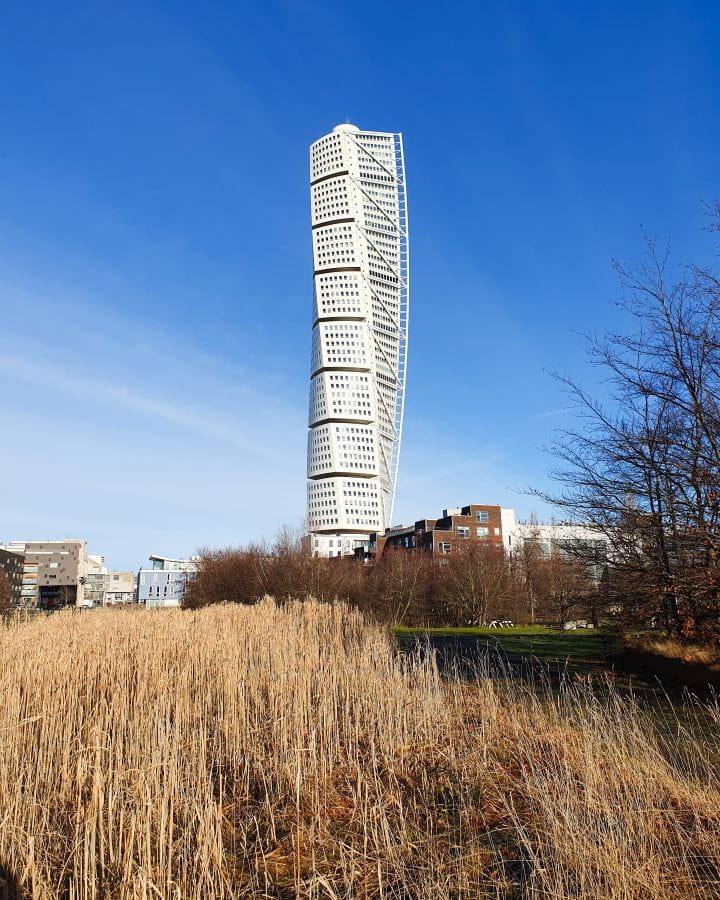 Turning Torso Sweden How To Have The Best Day In Malmö From Copenhagen In the foreground is brown dry grass and behind it is a tall white building that twists and has lots of windows on it in Malmö, Sweden. How To Have The Best Day In Malmö From Copenhagen