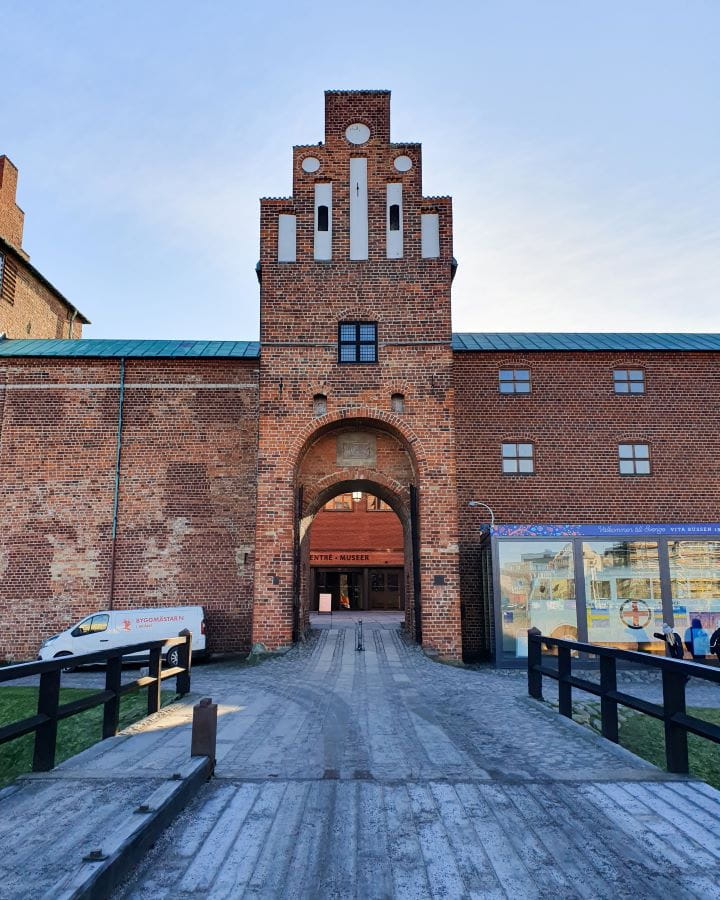 Malmö castle Sweden How To Have The Best Day In Malmö From Copenhagen A stone entryway to Malmö castle in Malmö, Sweden. The photo is taken on a wooden bridge leading into a stone archway with walls coming off it. There's little windows in the building and a white van out the front too. How To Have The Best Day In Malmö From Copenhagen