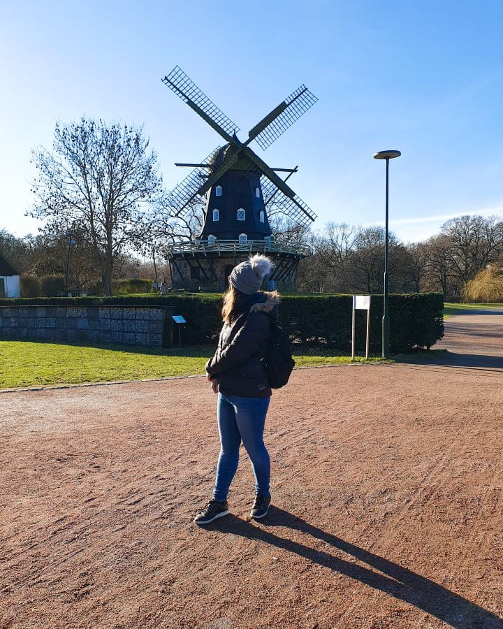 Sweden How To Have The Best Day In Malmö From Copenhagen Katherine standing on a dirt path with a small windmill behind her in Malmö, Sweden. The windmill has 4 blades and little windows on it and around it are trees and bushes. How To Have The Best Day In Malmö From Copenhagen
