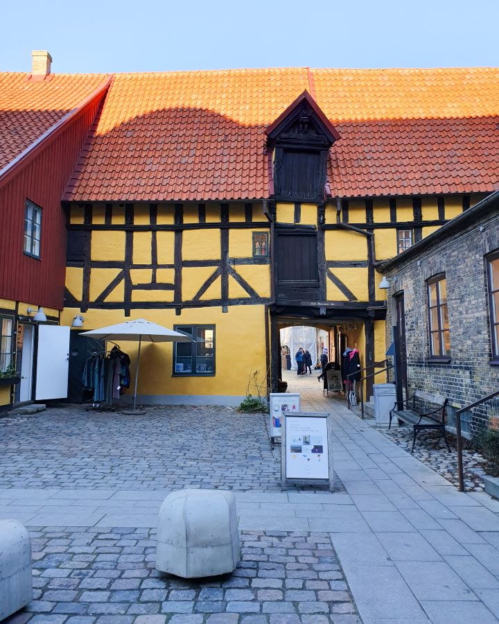 Casa Ekströmska Sweden How To Have The Best Day In Malmö From Copenhagen The old building Casa Ekströmska in Malmö, Sweden. The yellow building is wonky, with a red tiled roof and wooden details. There's a walkway through the building and a shop out front with an umbrella and some clothes on the cobblestones. How To Have The Best Day In Malmö From Copenhagen