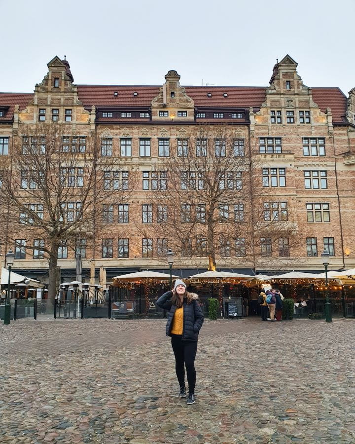 Stortorget Sweden How To Have The Best Day In Malmö From Copenhagen Katherine standing in a cobblestone square in Malmö, Sweden. Behind her is a beautiful European looking building with lots of windows and a brown roof. Under it are a bunch of restaurants with outdoor seating. How To Have The Best Day In Malmö From Copenhagen