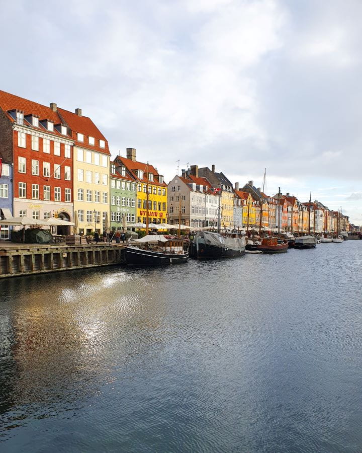 Nyhavn Denmark How to Enjoy Your Time in Copenhagen For Free Another view of Nyhavn in Copenhagen, Denmark. The canal has boats moored to the side and lots of coloured buildings going away from the camera beside it. There's seating on the side of the water and the sky overhead is cloudy. How to Enjoy Your Time in Copenhagen For Free