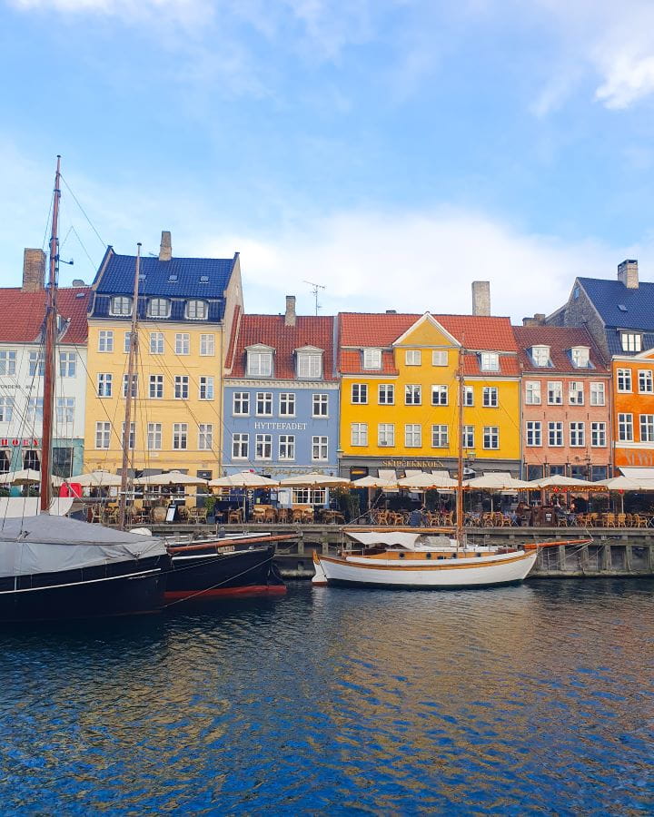 Nyhavn Denmark How to Enjoy Your Time in Copenhagen For Free How to Enjoy Your Time in Copenhagen For Free. Six coloured buildings in Copenhagen, Denmark. The buildings are yellow, orange, blue and white with orange and blue roofs and umbrellas and seating out the front. In the foreground is a canal with boats with masts in them and the sky is blue and cloudy.