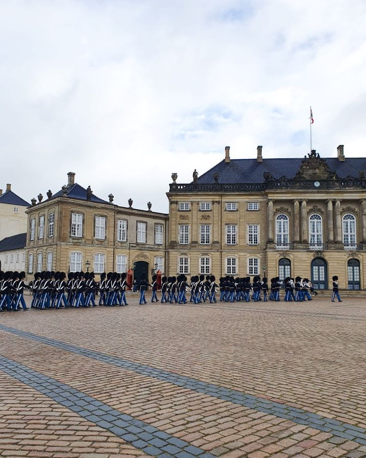Amalienborg Palace Denmark How to Enjoy Your Time in Copenhagen For Free A square in Copenhagen, Denmark made of bricks. There's lots of guards with blue pants, and fluffy black hats walking in lines. Behind them is one of the palaces with a blue roof and big white windows. How to Enjoy Your Time in Copenhagen For Free