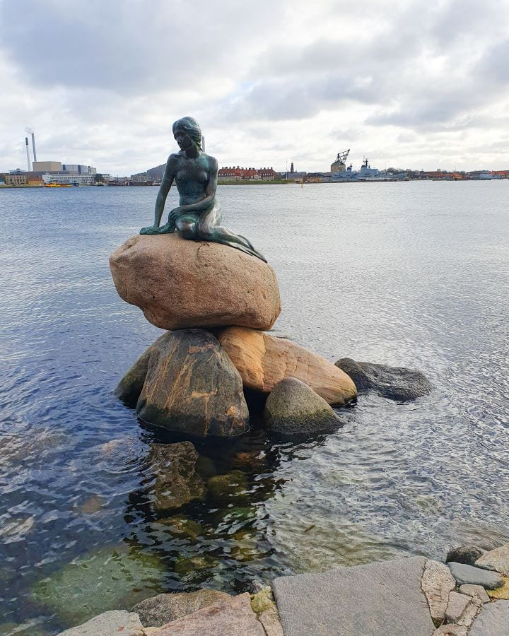 Little Mermaid Statue Denmark How to Enjoy Your Time in Copenhagen For Free A bronze statue of a woman mermaid sitting on a couple of rocks in the middle of the water in Copenhagen, Denmark. Behind the water on the land is lots of factories and buildings and the sky is overcast. How to Enjoy Your Time in Copenhagen For Free