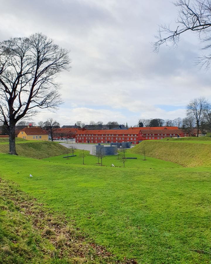 Kastellet Fortress Denmark How to Enjoy Your Time in Copenhagen For Free Inside the Kastellet Fortress in Copenhagen, Denmark which has lots of short green manicured grass with bare trees on the side and orange buildings in the distance. How to Enjoy Your Time in Copenhagen For Free
