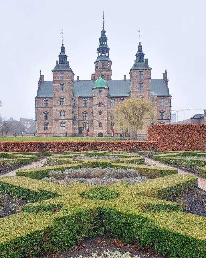 King's Garden Rosenborg Castle Denmark How to Enjoy Your Time in Copenhagen For Free Short green hedges in shapes with paths between them and a neat orange hedge lining the garden. Behind it is Rosenborg Castle which is made of stone with a grey roof and 4 turrets with windows all over it in Copenhagen, Denmark. How to Enjoy Your Time in Copenhagen For Free