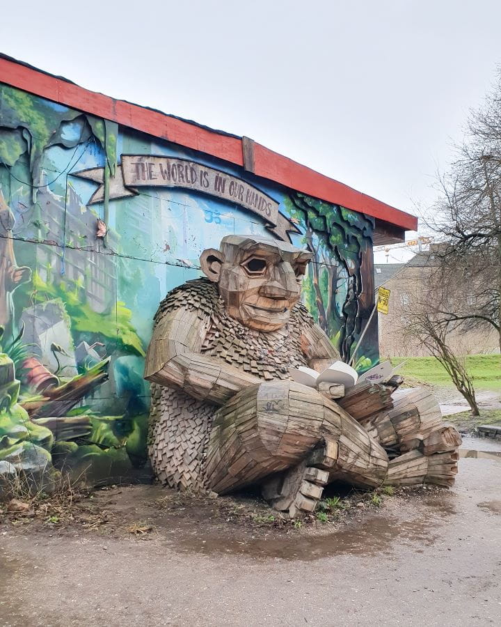 Freetown Christiania Denmark How to Enjoy Your Time in Copenhagen For Free A big wooden troll statue in Freetown Christiania in Copenhagen, Denmark. It's sitting cross legged in front of a nature green mural with the words "The world is in our hands" above it. How to Enjoy Your Time in Copenhagen For Free