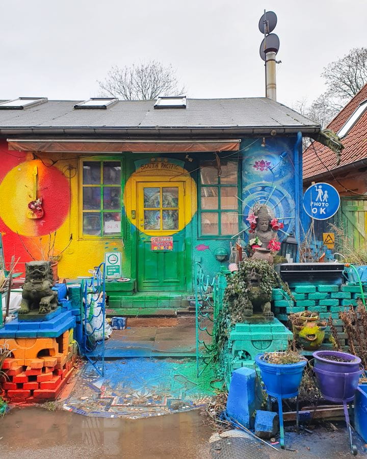 Freetown Christiania Denmark How to Enjoy Your Time in Copenhagen For Free A little house in Freetown Christiania in Copenhagen, Denmark painted green, blue, red and yellow. Out front there's coloured bricks. pot plants and some statues and plants. How to Enjoy Your Time in Copenhagen For Free