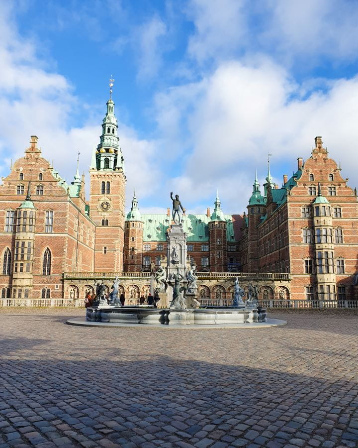 Denmark Explore Frederiksborg Castle In a Beautiful Day Trip from Copenhagen A cobblestone square with a big fountain in the middle. Behind it is a big brown castle with turrets and a green roof in Denmark. Explore Frederiksborg Castle In a Beautiful Day Trip from Copenhagen