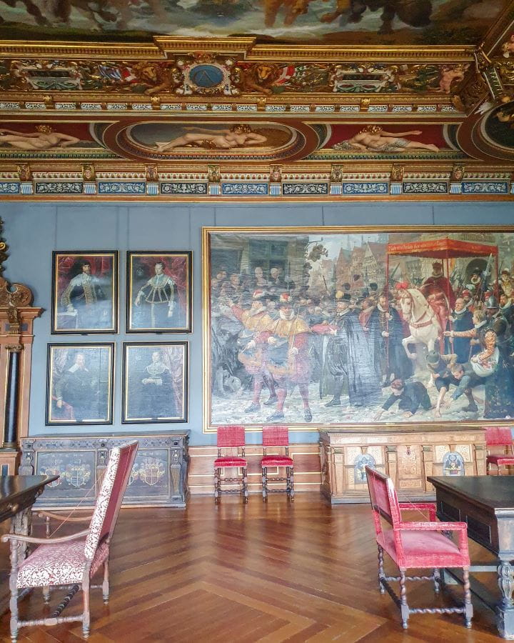 Denmark Explore Frederiksborg Castle In a Beautiful Day Trip from Copenhagen Inside a room in Frederiksborg castle in Denmark. The floor is wooden and there are 5 red chairs with some wooden tables. There's some other furniture and on the walls are 5 paintings. The roof is gold and red and also painted and is very grand. Explore Frederiksborg Castle In a Beautiful Day Trip from Copenhagen