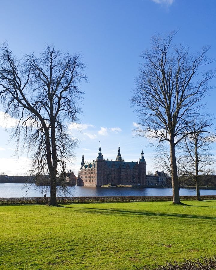 Denmark Explore Frederiksborg Castle In a Beautiful Day Trip from Copenhagen Neat green grass with big bare trees framing the view of the castle on the other side of the pond in Denmark. The sky and water are blue and the castle is slightly silhouetted. Explore Frederiksborg Castle In a Beautiful Day Trip from Copenhagen